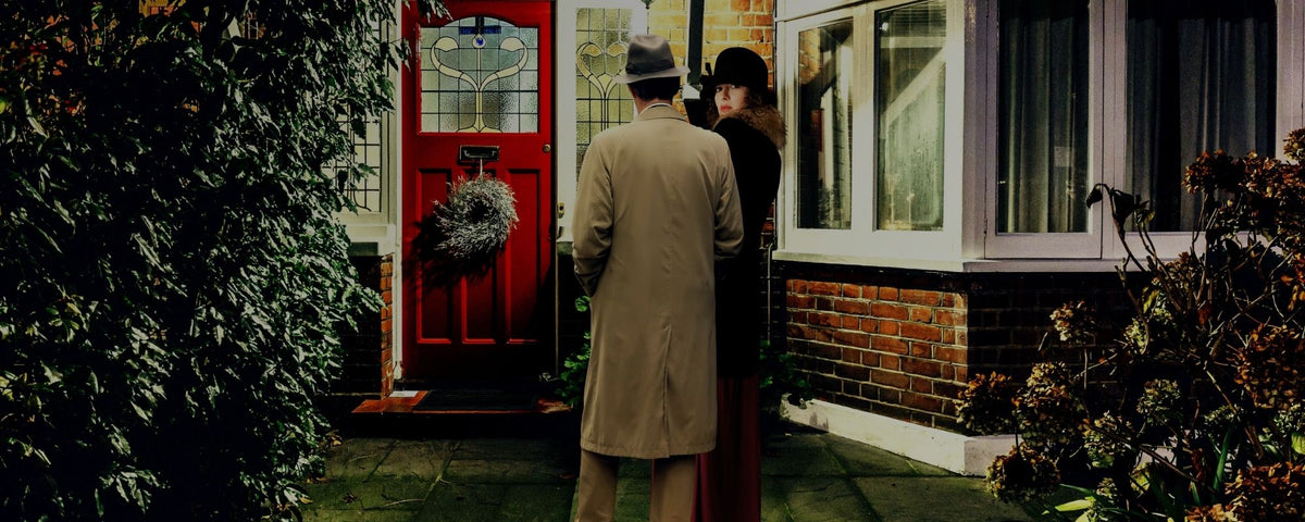 A man in a beige trench coat and a woman in a dark dress stand in front of a red front door of a brick house at night.