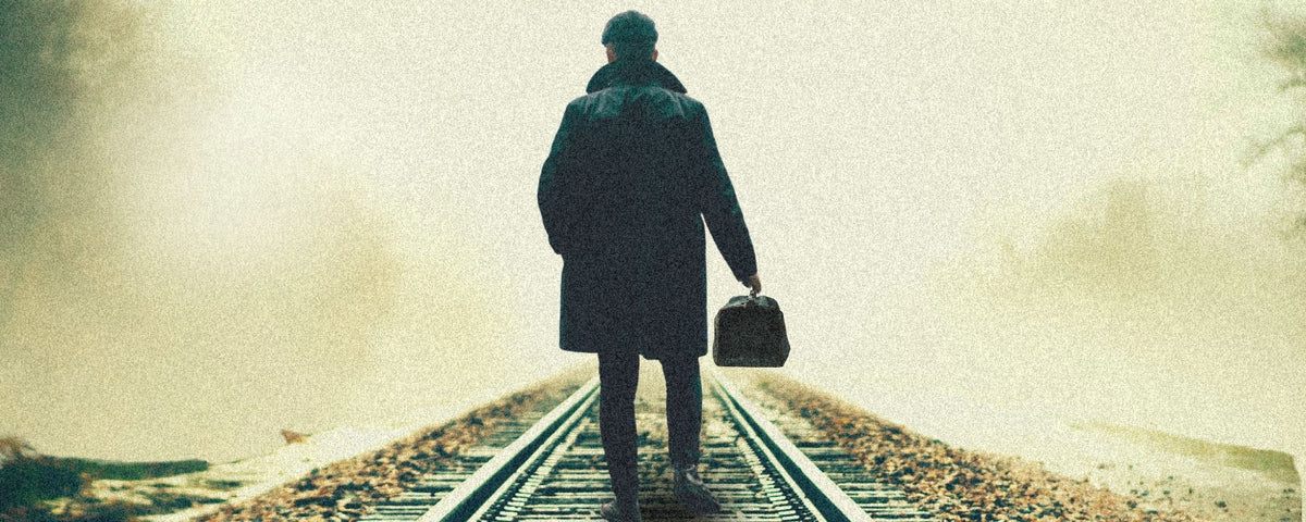 A person seen from behind walking away on railroad tracks, wearing a dark coat and carrying a small bag, with a hazy, sepia-toned outdoor background.