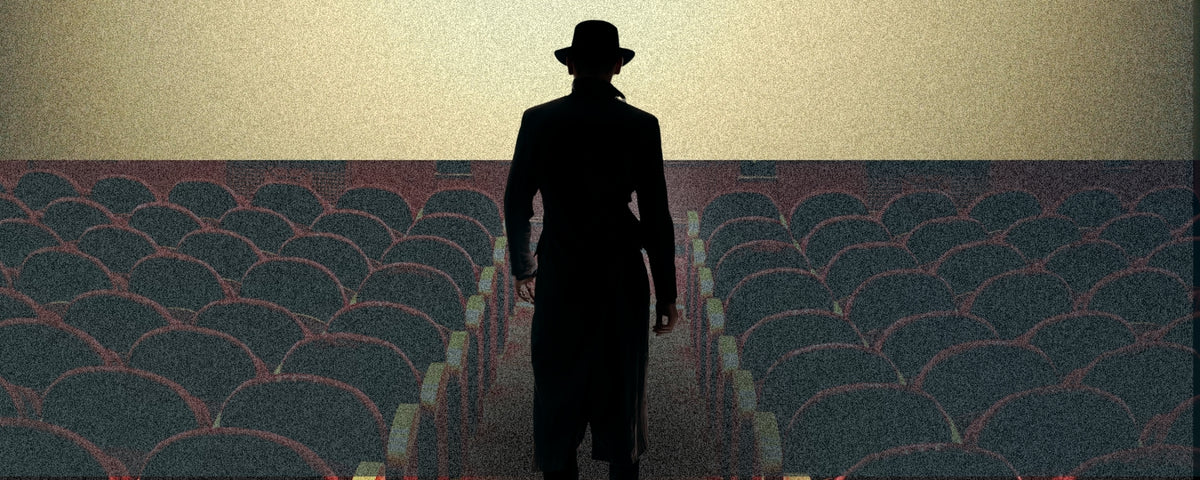 A silhouetted person wearing a fedora and long coat stands in the center aisle between rows of dark theater seats under a pale sky.